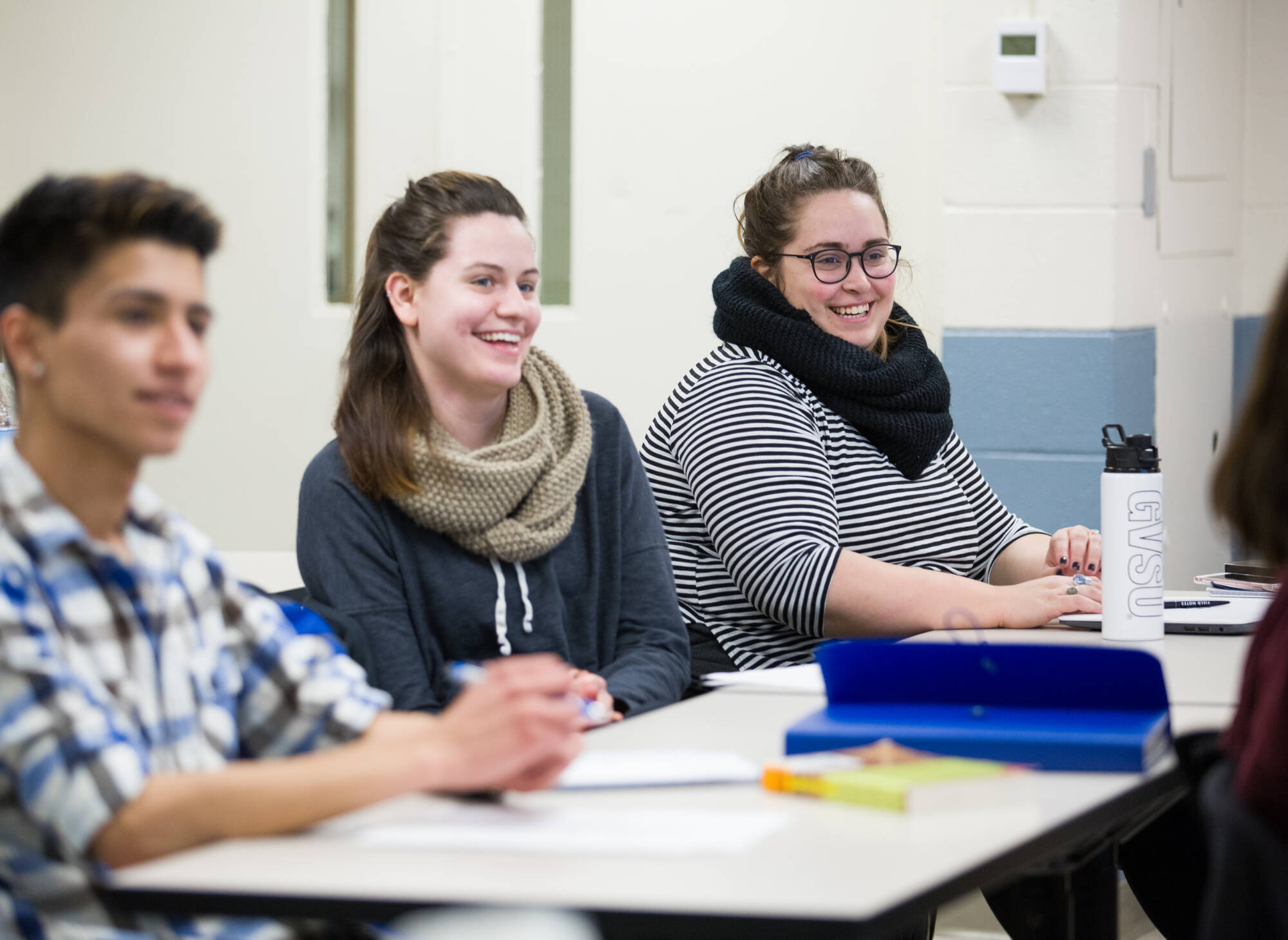 Students seated at classroom tables smile and engage in discussion, with notebooks and materials in front of them.